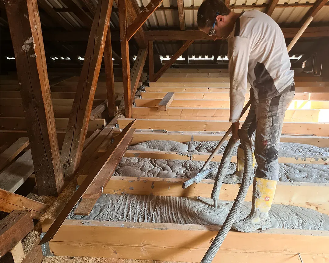 Ein Dachboden mit Holzbalken, die mit frischer Schüttdämmung befüllt werden, ein Arbeiter mit einem Werkzeug in Aktion